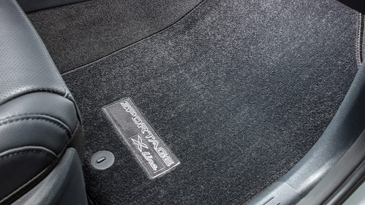  Close-up view of the 2027 Kia Sportage Hybrid SUV's front passenger‑side floor area, featuring a dark carpeted floor mat with stitched X-Line branding.