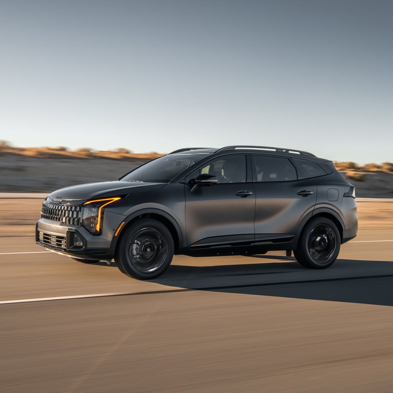 A gray 2027 Kia Sportage Plug-In Hybrid SUV speeding along a desert highway, highlighting the driver’s side, sleek design, LED amber daytime lights, and all-wheel drive.