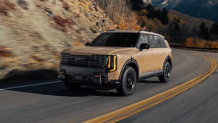  A dynamic action shot of the 2027 next-generation Kia Telluride in tan driving on a winding road, featuring rugged off-road design and black accents. The SUV is surrounded by mountains and fall foliage.