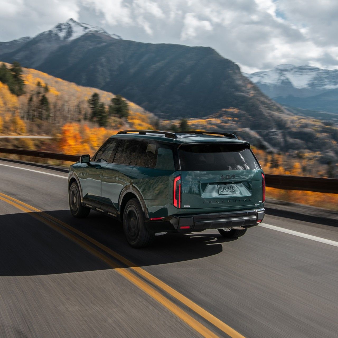 An action shot of the 2027 Kia Telluride Hybrid X-Line SX Prestige in blue-green driving on a highway, with a dense forest and snowy mountains in the background. The image highlights the rear and driver-side view of the SUV.