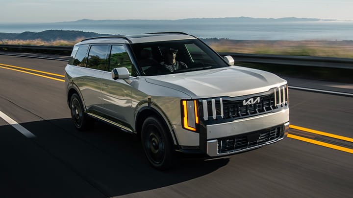 Foto profesional de un Telluride Hybrid SUV 2027 en color gris conduciendo rápidamente sobre una carretera lisa, con vegetación y colinas envueltas en niebla al fondo.