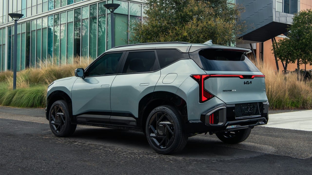 2027 Kia Seltos Hybrid en azul, vista trasera de tres cuartos estacionado en una carretera con árboles y edificios modernos al fondo, destacando sus elegantes luces traseras LED, diseño del techo y manijas de puerta automáticas.