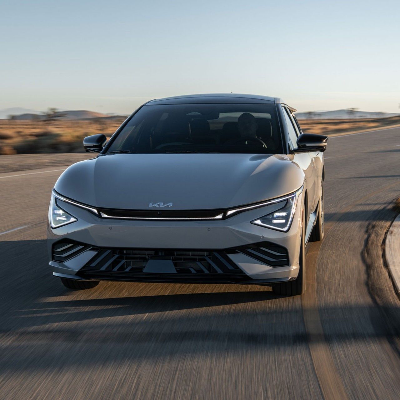 Una emocionante toma de acción del Kia EV6 Wind AWD 2026 en color gris mientras se dirige hacia el espectador, descendiendo por una carretera curva en el desierto con arena y arbustos visibles en el fondo. El CUV luce elegante y dinámico con su diseño frontal y faros LED.