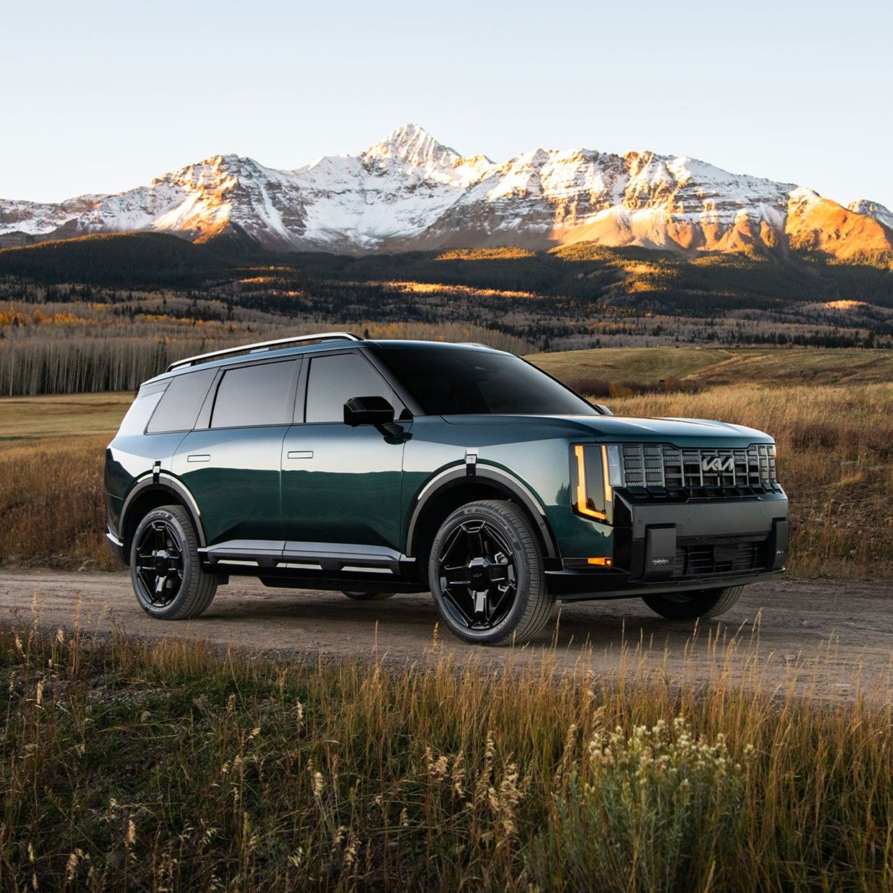 Una imagen fija del Kia Telluride Hybrid 2027 en un exterior azul-verde, posicionado en un camino de tierra con el lado del pasajero frente a la cámara. El SUV está rodeado de plantas y se encuentra frente a picos montañosos nevados.